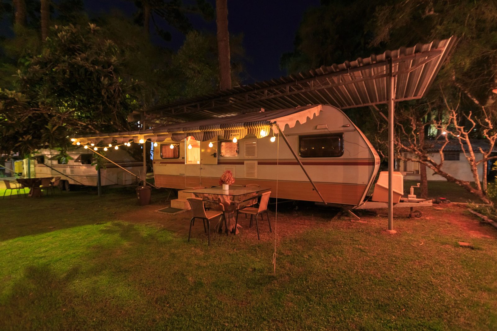 Camper at dusk with the awning out, string lights on, and a table and chairs set up outside