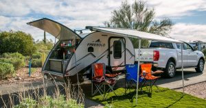 Teardrop camper parked next to a truck with the awning out, camp chairs and an outdoor rug set up
