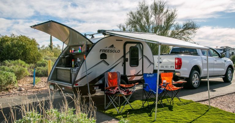 Teardrop camper parked next to a truck with the awning out, camp chairs and an outdoor rug set up