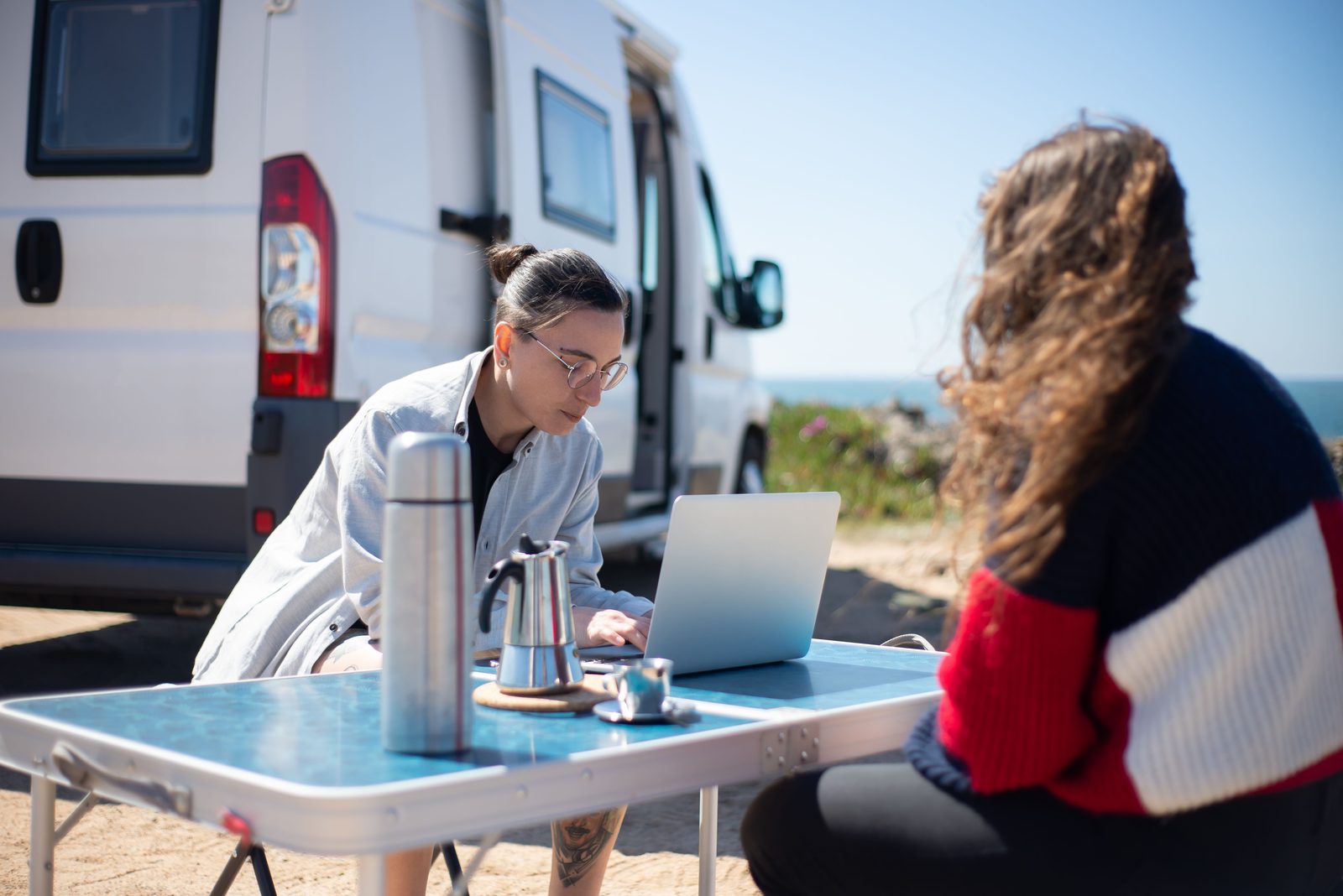 Camper working on a laptop at a folding table outside a van with a thermos and coffee pot
