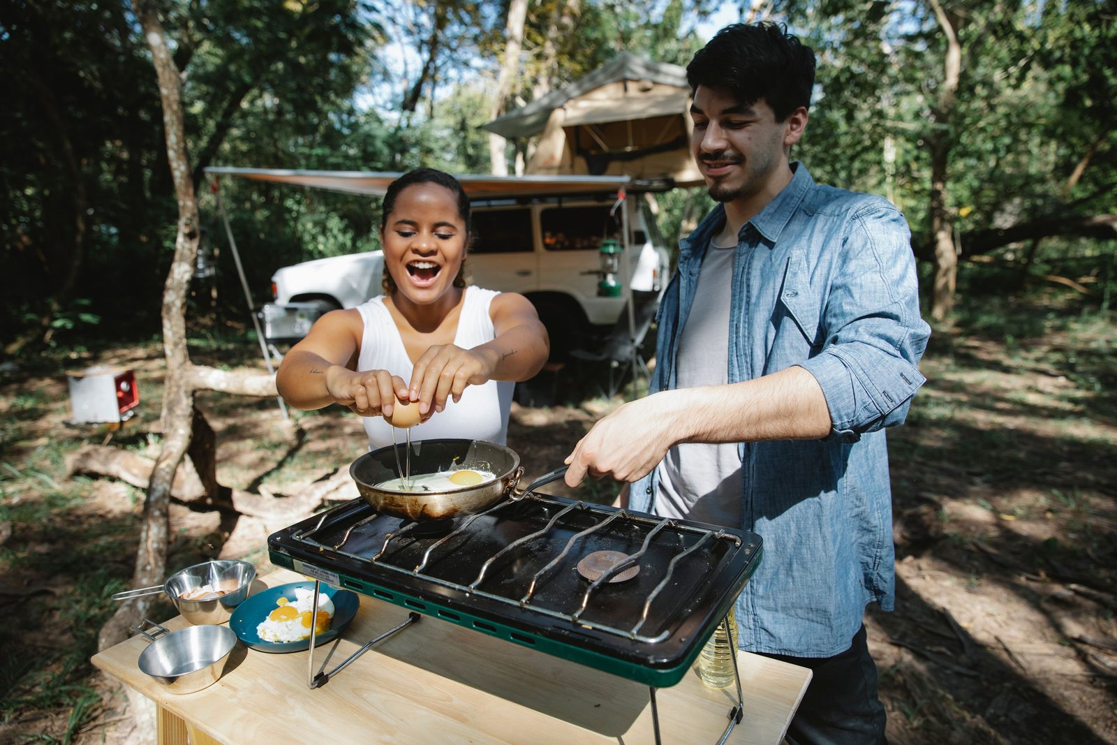 Two campers cooking breakfast on a camp stove set up on a folding table outside a camper