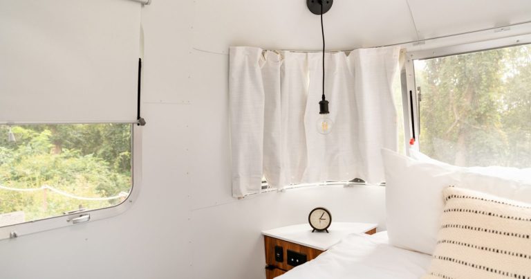 Bright white camper bedroom interior with a made bed, curved RV windows and blank walls ready for wallpaper